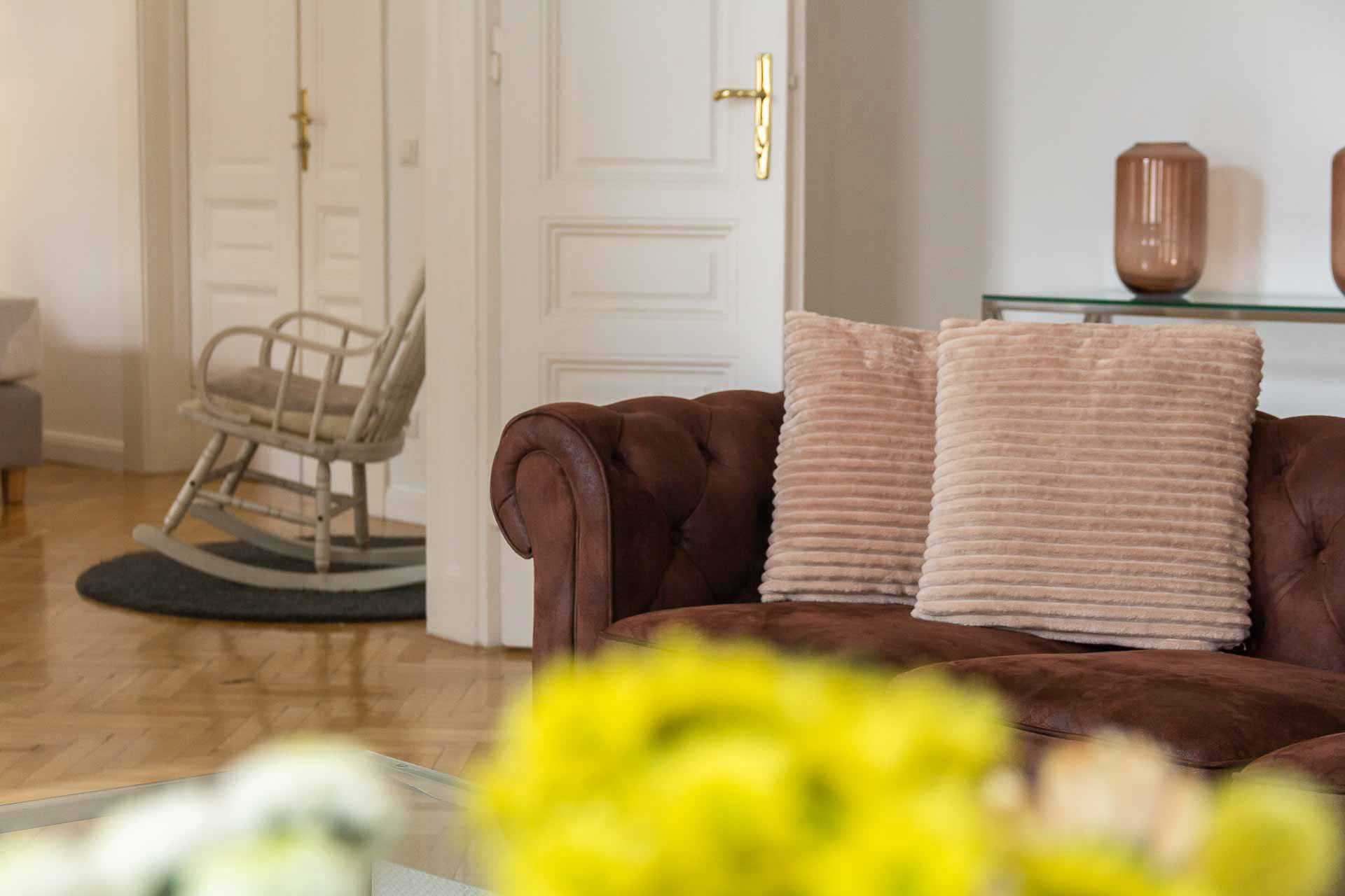 Fresh flowers and an elegant brown couch with light brown cozy cushions on it; a rocking chair in the back