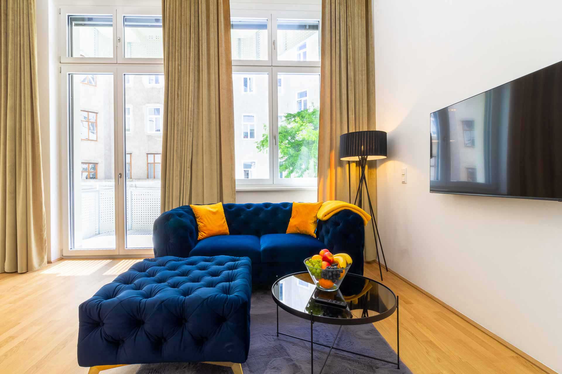 Big windows with beige curtains, in front a luxuroius blue velvet couch with yellow cushions on it, in Viennas 5th district