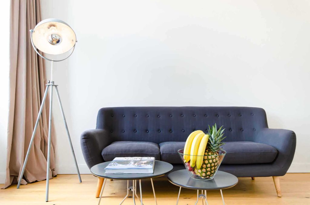 Front view if a cozy modern couch with two endtables with magazines and fresh fruit on it, on the left side is a round and modern lamp with a beige curtain in the back, located in Vienna