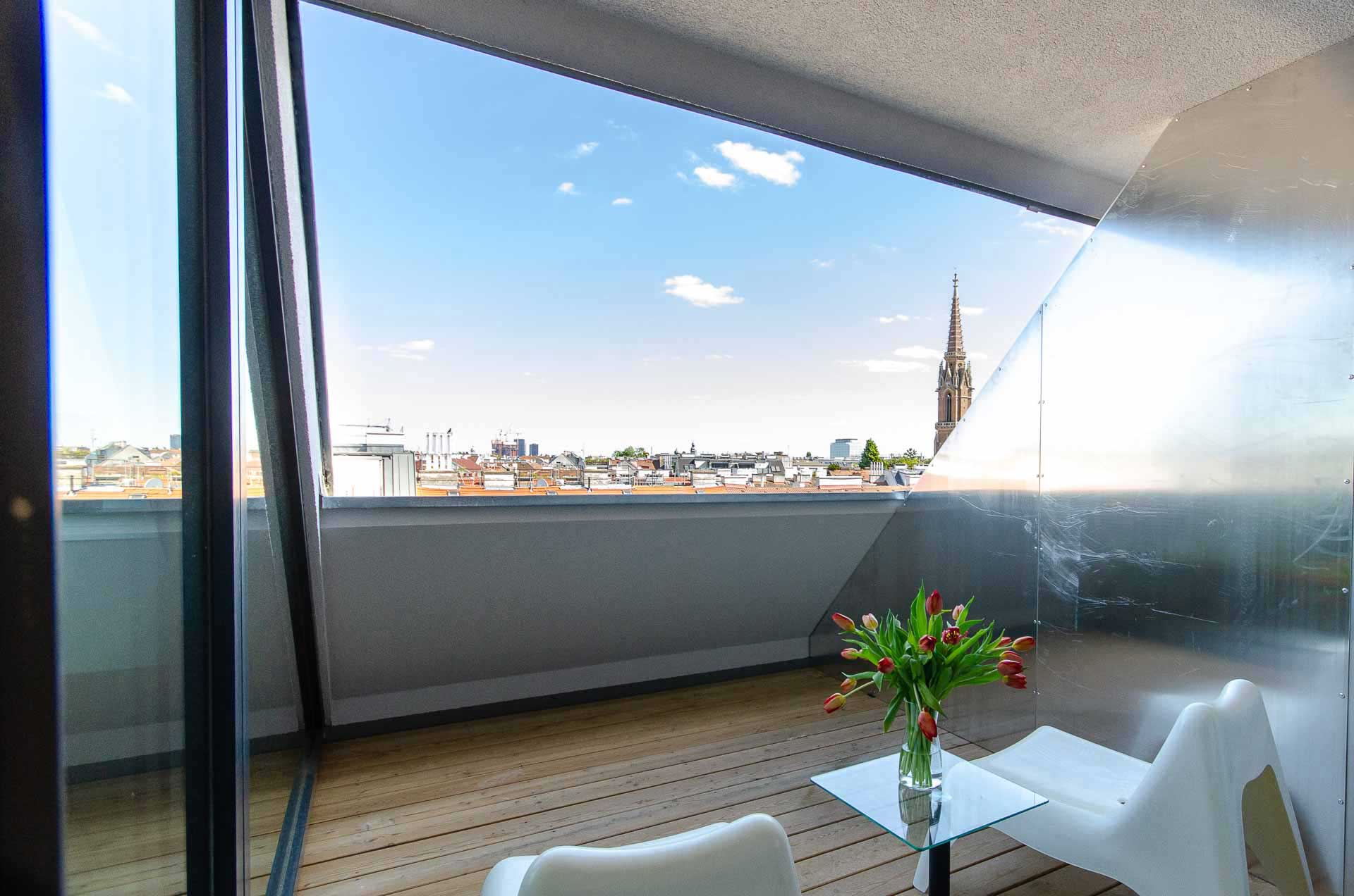 Balcony with wooden floors and two white lounge chairs on it, in the middel a glass table with fresh tulips on it; beautiful view of vienna and blue sky