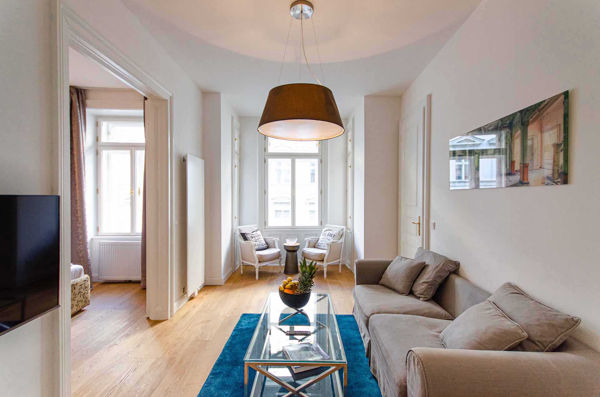 Living room with cozy grey couch, glass table and blue carpet; in the back two cozy lounge chairs
