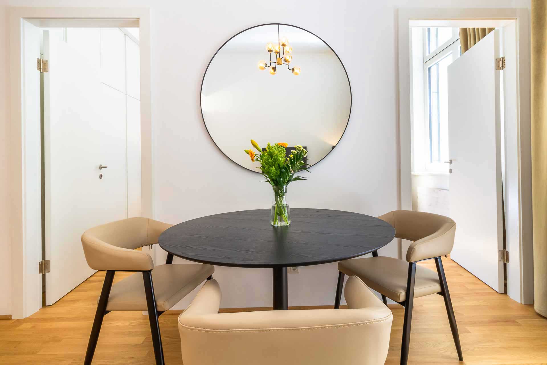 View of a dining area with a black round table with fresh floewers on it; and three beige leather chairs and a round mirror on the wall