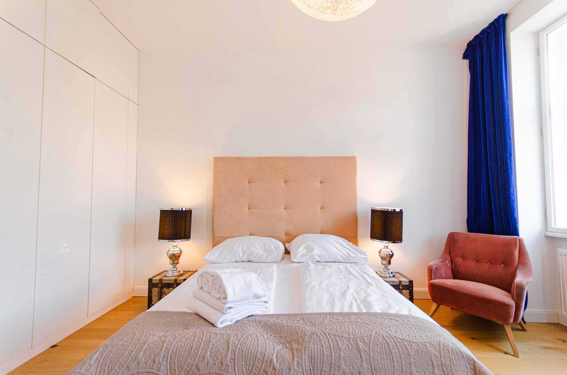 Modern and cozy bedroom with a beige velvet kingsize bed with fresh linen and towels; on the right a red velevet chair and blue curtains; on the left a big white built in wardrobe