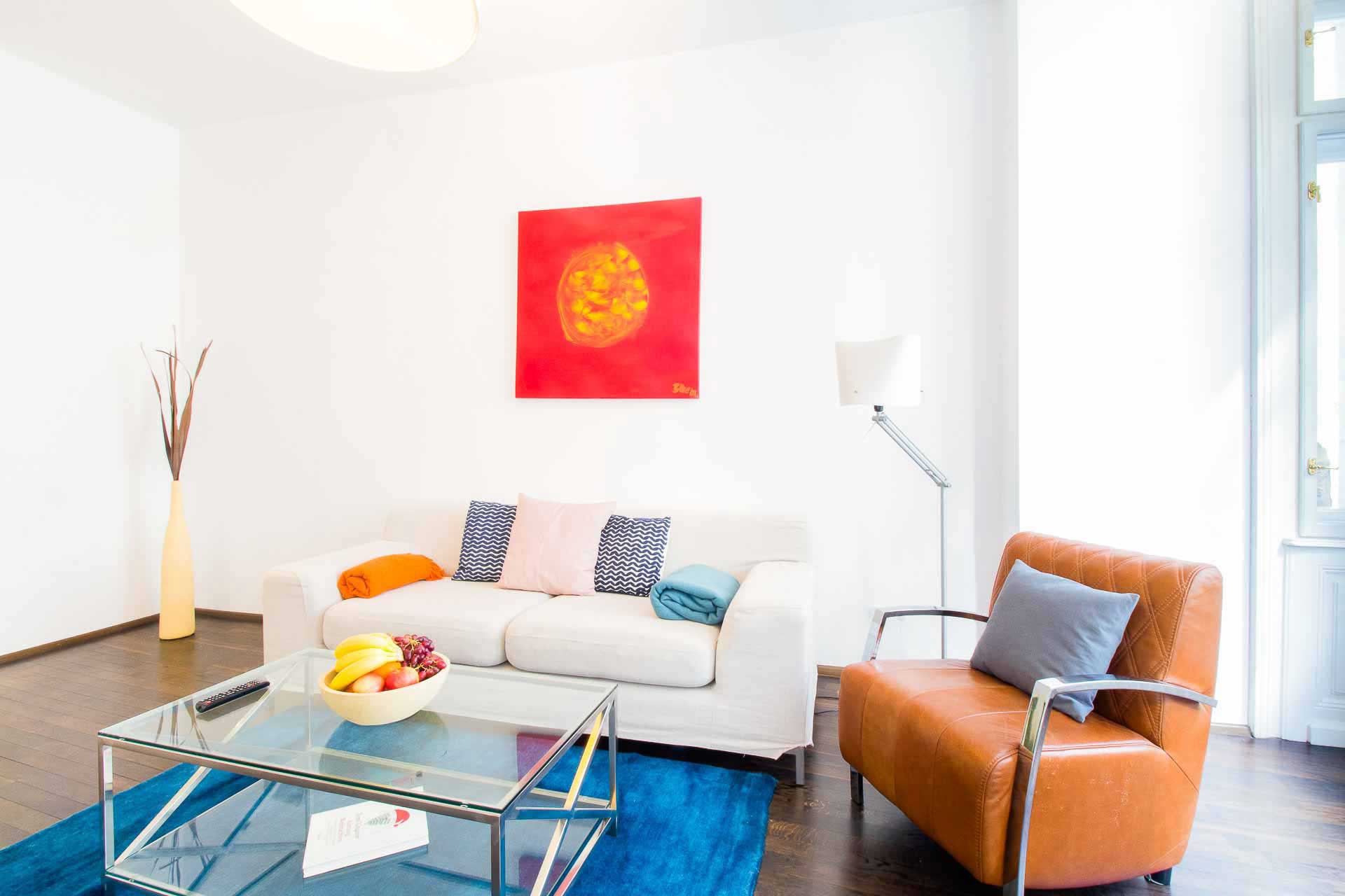 View of a living room: red paining on the wall, white couch with cozy cushions on it, on the right side a brown leather chair, in front of it a glass end table and a blue carpet
