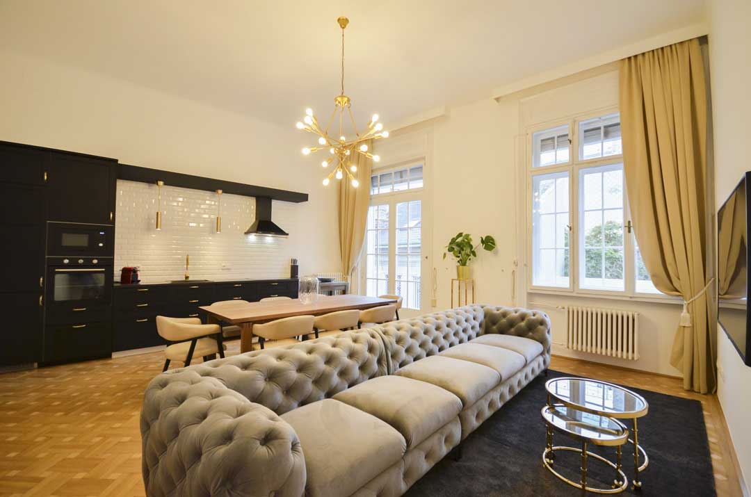 In the back: elegant black kitchen and a long wooden table; in the front: a cozy grey velvet couch and a golden end table