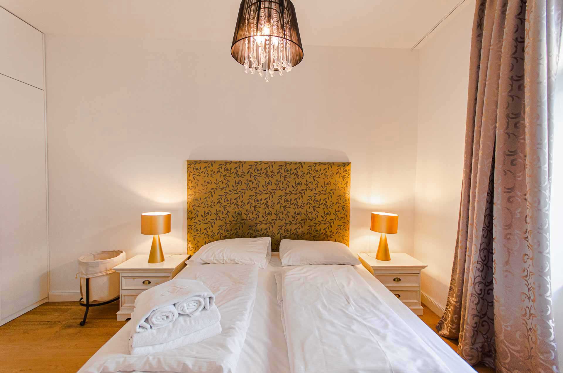 Golden kingsize bed with fresh linen and towels, two white end tables on the left and right side with golden lamps on them