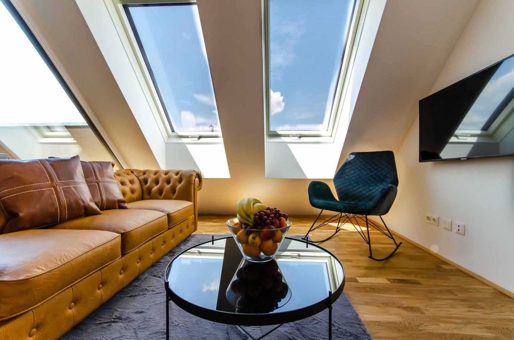 A cozy living room in a vacation rental featuring a brown leather couch, a rug beneath a coffee table with a bowl of fresh fruit on top. An armchair sits in the corner, and two large windows above the table fill this penthouse space with natural light.